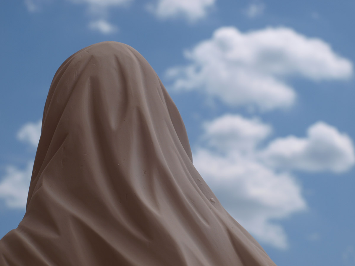 Im Hintergrund ist Wolkenhimmel zu sehen, im Vordergrund eine Madonnenfigur von hinten.